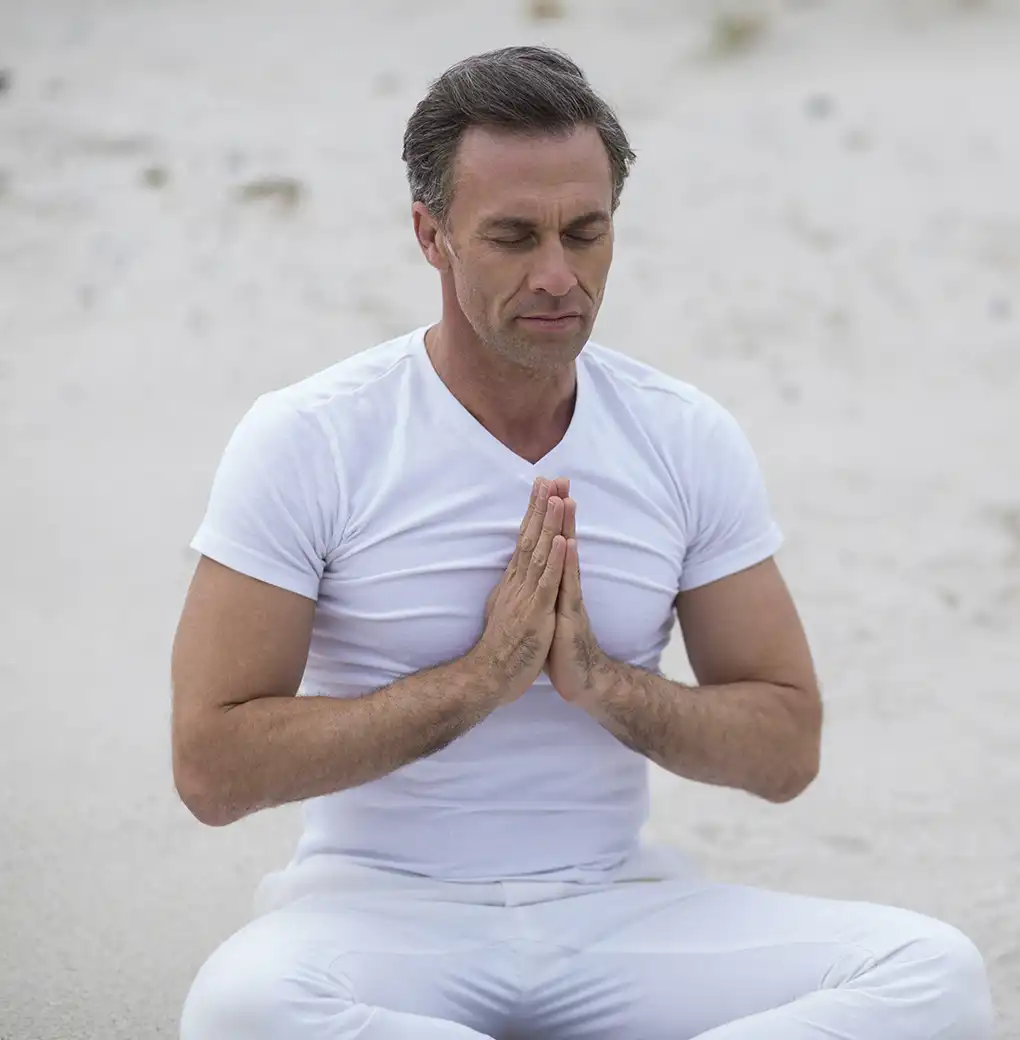Hombre meditando en la playa con las manos en posición de oración, practicando una visualización de amor propio para equilibrar su energía interior